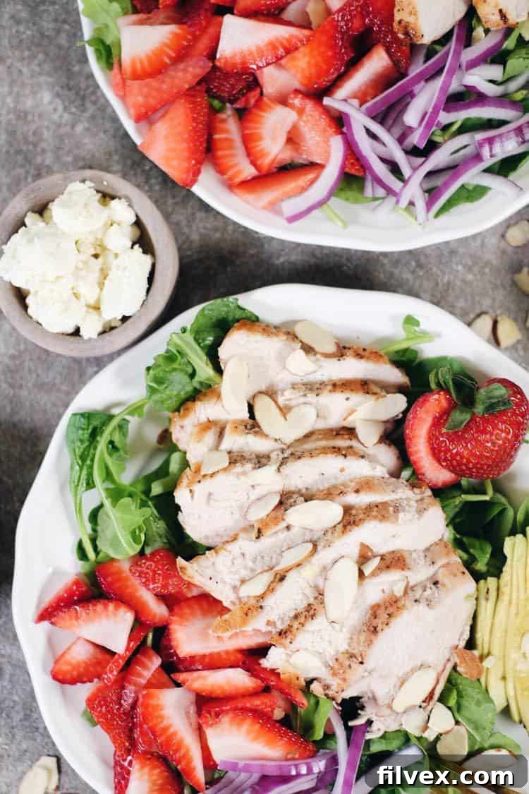 A vibrant bowl of fresh strawberry chicken salad featuring a mix of arugula and spinach, sliced red strawberries, creamy avocado, and red onion, all topped with crunchy almonds and ready for dressing.
