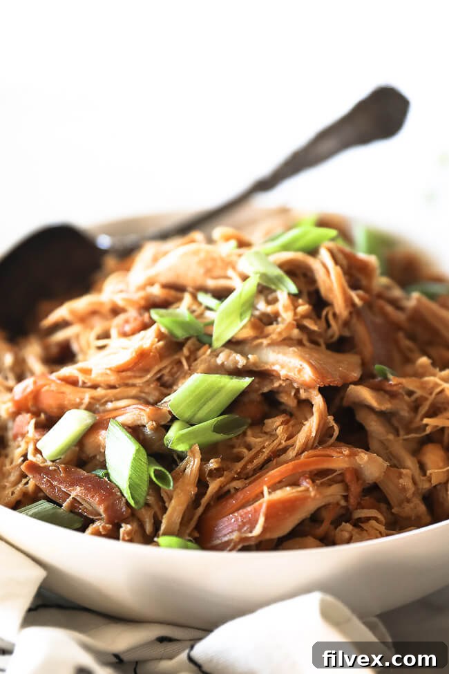 Angled close up image of instant pot honey garlic chicken in bowl with green onion on top and serving spoon dug into bowl in the background. 