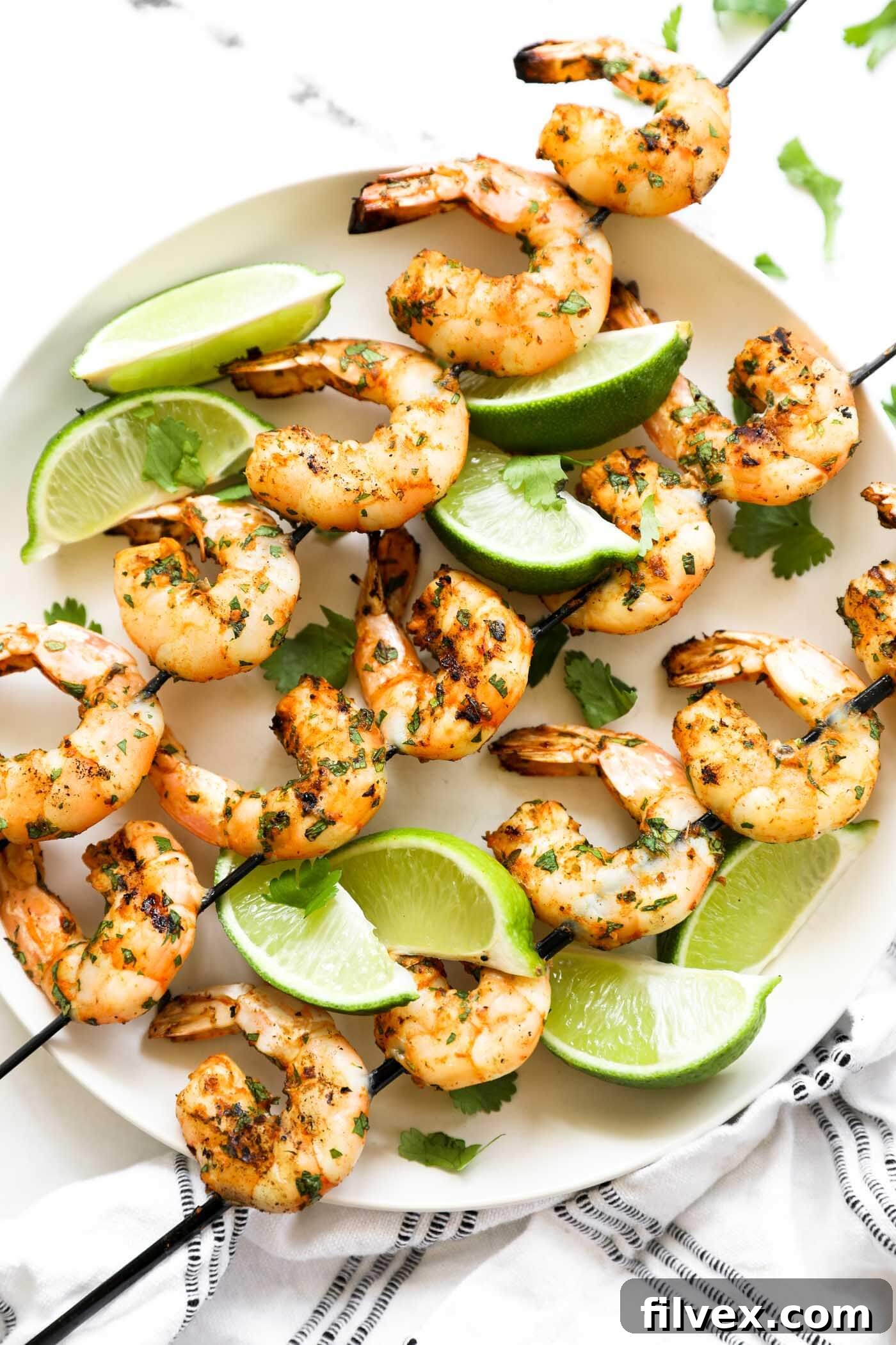 Grilled shrimp kabobs laid out on a plate. 3 skewers with lime wedges and fresh cilantro garnish.
