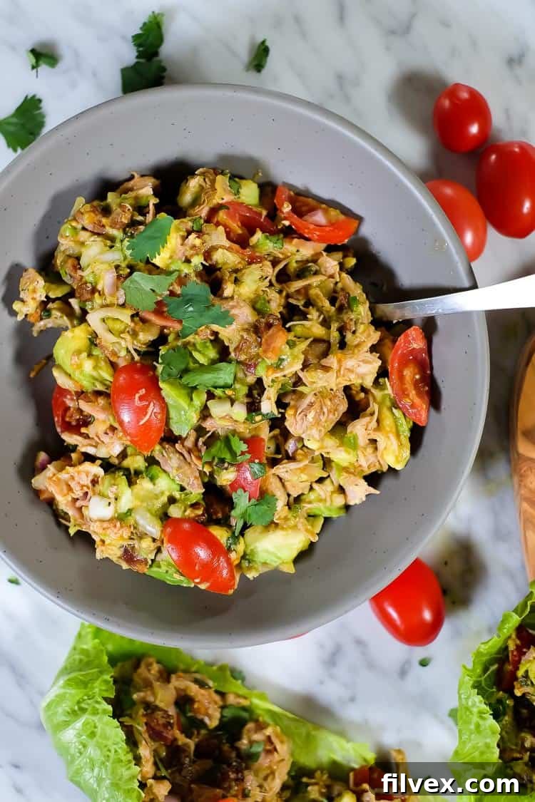 Close-up of a spoon scooping creamy Paleo chicken avocado salad with visible chunks of avocado, shredded chicken, and crispy bacon. Fresh red onion and cilantro add color and texture.