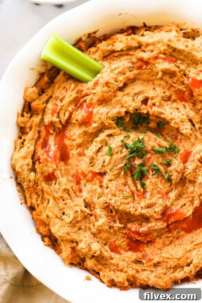 Close up overhead image of a round baking dish with creamy dairy-free buffalo chicken dip. A sprinkle of hot sauce on top, garnished with fresh chopped parsley and a celery stick dug into the dip, highlighting its vibrant texture and inviting presentation.