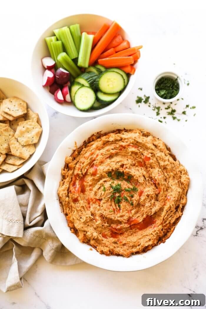 Overhead image of a round dish filled with dairy-free buffalo chicken dip, surrounded by bowls of fresh sliced vegetables like celery and carrots, and gluten-free crackers on a wooden serving board for dipping. This wholesome spread is presented attractively.