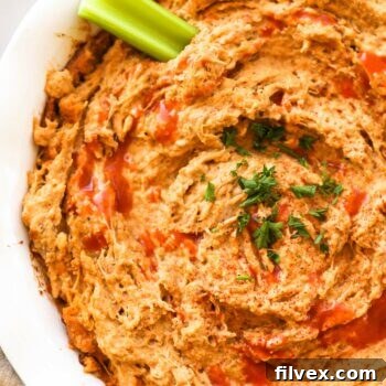 Close up overhead image of a round baking dish with creamy dairy-free buffalo chicken dip. A little extra hot sauce sprinkled on top, garnished with chopped parsley and a celery stick dug into the dip.