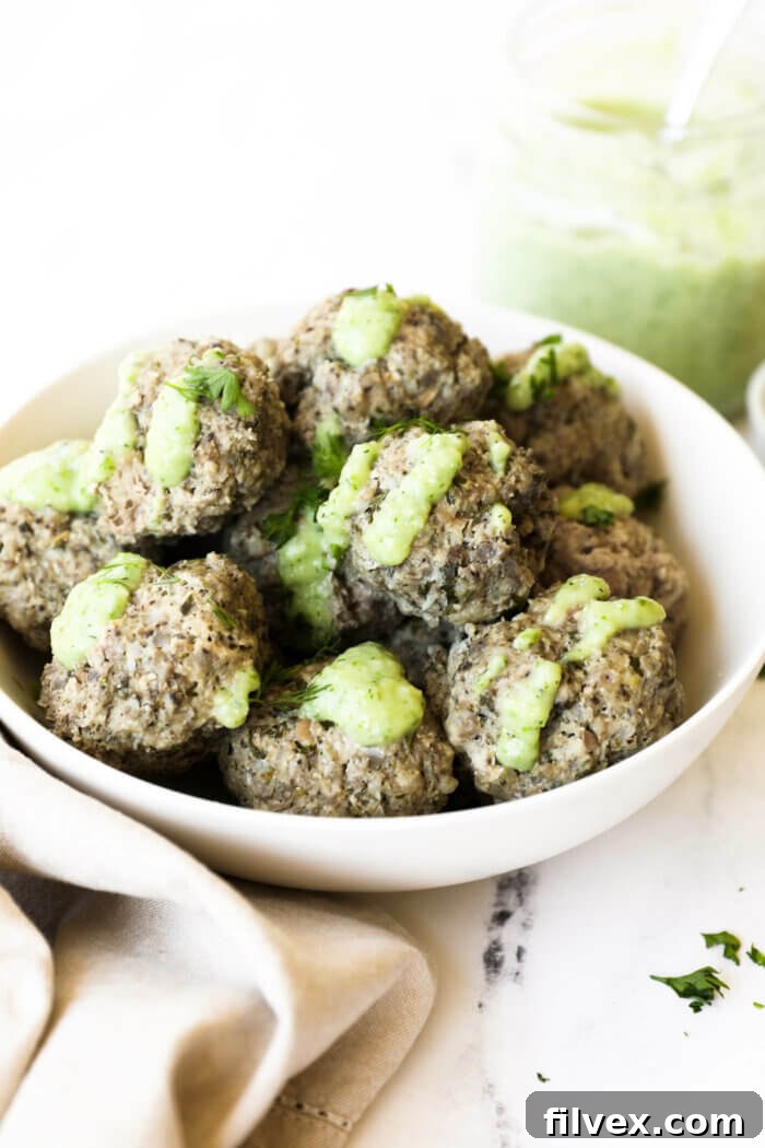 A large platter with various Greek meatballs, some sliced, served with a bowl of dairy-free tzatziki, fresh dill, and lemon wedges, showcasing serving options.