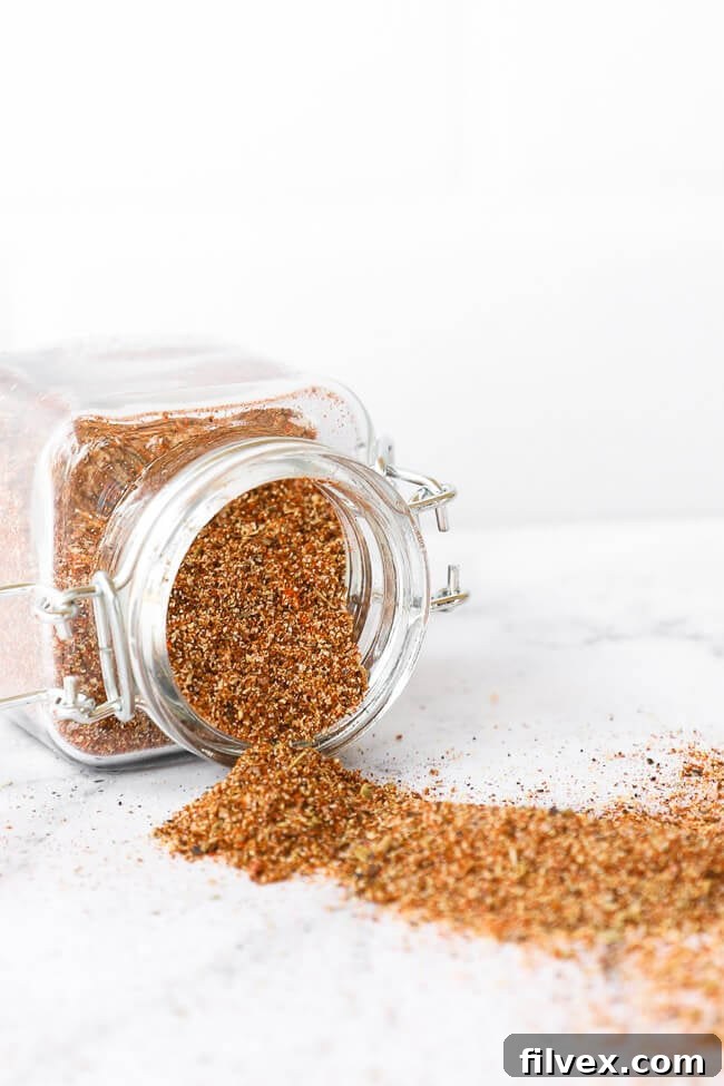 A glass jar of homemade taco seasoning tipped on its side, with a vibrant blend of spices spilling out onto a wooden surface