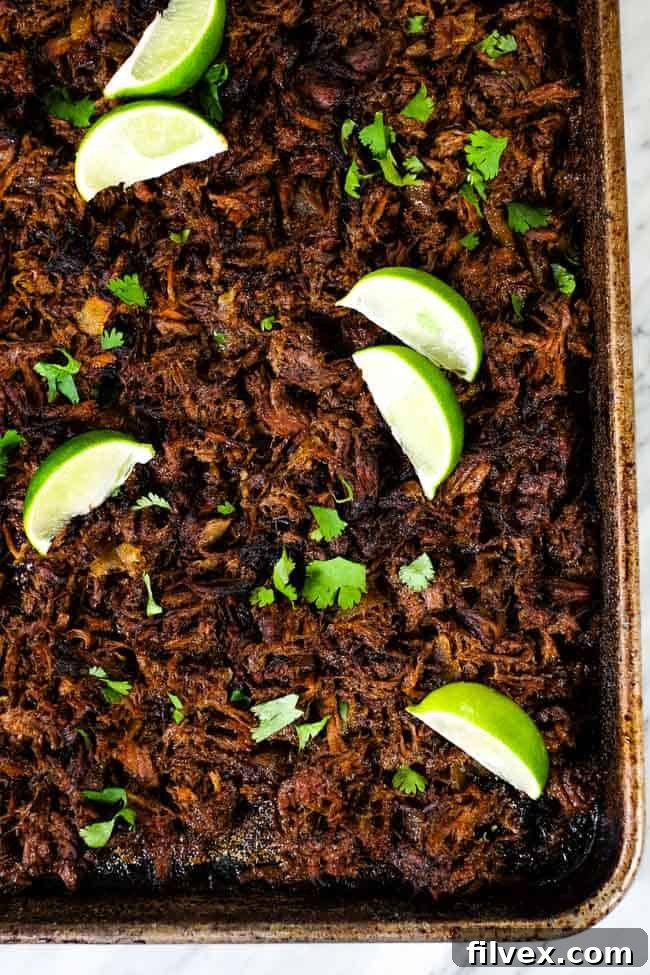 An inviting overhead vertical image of rich, shredded beef barbacoa spread on a sheet pan, artfully topped with fresh cilantro and vibrant lime wedges, ready to be served or crisped.