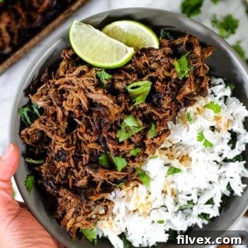 Overhead image of a hand holding a bowl of greens, white rice and shredded beef garnished with chopped cilantro and lime wedges, emphasizing a delicious, ready-to-eat meal.