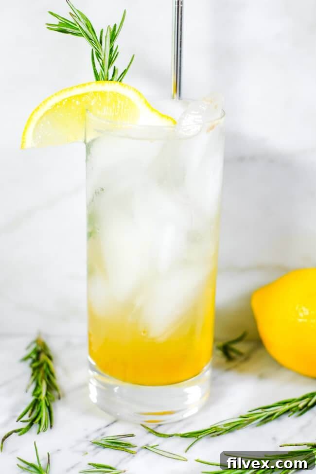Tall glass of iced lemon & rosemary cocktail with lemon wedge and fresh rosemary garnish. 