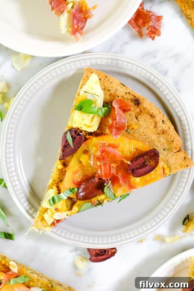 A slice of grilled paleo pizza, adorned with heirloom tomatoes, artichoke hearts, kalamata olives, prosciutto, and fresh basil, resting on a plate.