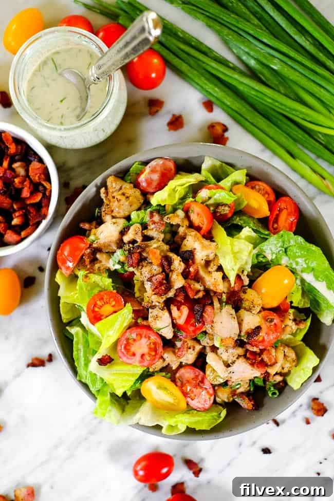 A close-up view of a BLT salad in a clear bowl, showcasing diced tomatoes, crispy bacon, and a creamy dressing drizzle. A sprig of green onion provides a fresh garnish.