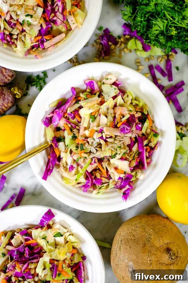 Three bowls of vibrant Paleo and Whole30 coleslaw are arranged on a rustic wooden table, surrounded by the fresh ingredients used in the recipe: whole lemons, bunches of fresh parsley, and various forms of cabbage and carrots. The scene emphasizes the simplicity and freshness of this easy, mayo-free coleslaw recipe.