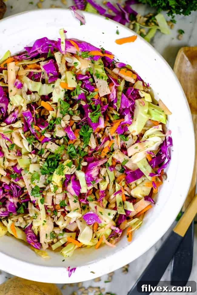 A large serving bowl filled with the colorful Paleo and Whole30 coleslaw is placed on a wooden table, with serving utensils ready. Around the bowl, individual components of the salad – thinly sliced jicama, shredded carrots, and various cabbages – are scattered, inviting viewers to appreciate the fresh ingredients that make up this easy and healthy side dish.