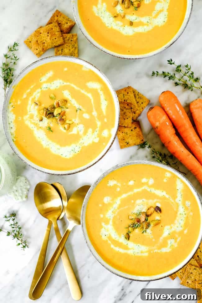 Three bowls of carrot ginger soup, beautifully topped with dairy-free tzatziki, crushed pistachios, and fresh thyme, artfully arranged on a wooden table alongside fresh ingredients and crackers.