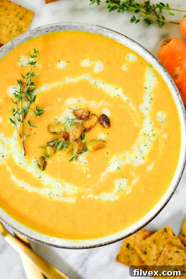 A close-up view of carrot ginger soup, highlighting the creamy texture and the elegant swirl of dairy-free tzatziki, pistachios, and a sprig of fresh thyme.