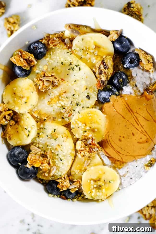 Close-up shot of a single serving of Chai Spiced Paleo Chia Seed Pudding, generously adorned with fresh banana slices, crisp pear wedges, bright blueberries, crunchy grain-free granola, and a decadent drizzle of nut butter.