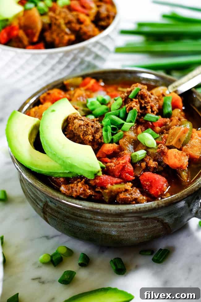 Angled shot of Paleo + Whole30 sweet potato chili in a bowl with chopped green onion and avocado slices, showcasing the inviting presentation and rich texture.