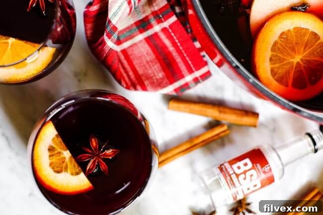 Simple mulled wine recipe in glasses with orange slices, cinnamon sticks, a plaid napkin, star anise and bourbon bottle. 