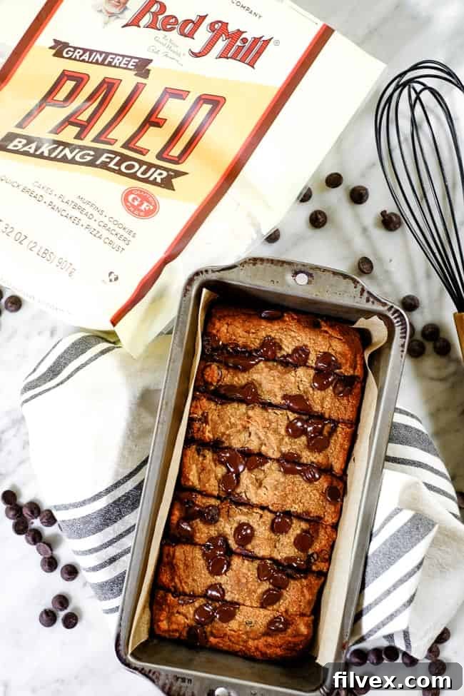 Ingredients for Paleo chocolate chip banana bread including Bob's Red Mill Paleo Baking Flour, chocolate chips, and a whisk, arranged on a rustic table next to a baked loaf.