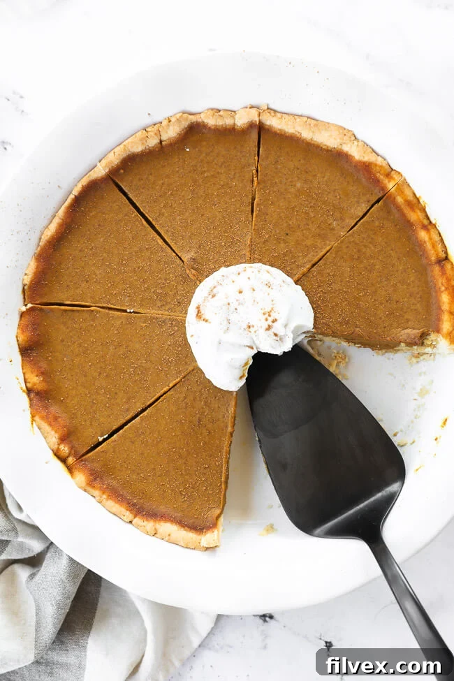 A stunning overhead shot of the dairy-free and gluten-free pumpkin pie in its dish, with two slices already removed. A pie server rests in the dish, alongside a dollop of whipped coconut cream and a dusting of cinnamon, inviting guests to serve themselves.