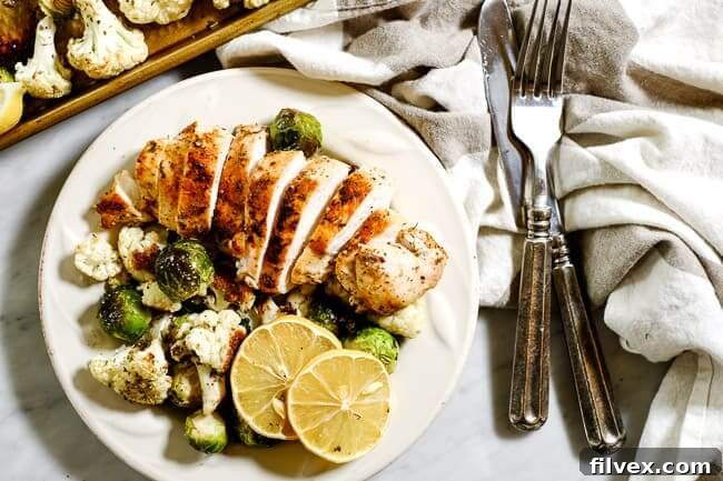 Easy lemon chicken served on a plate with roasted brussels sprouts and cauliflower and lemon slices for garnish, a grey and white striped napkin and silverware. 