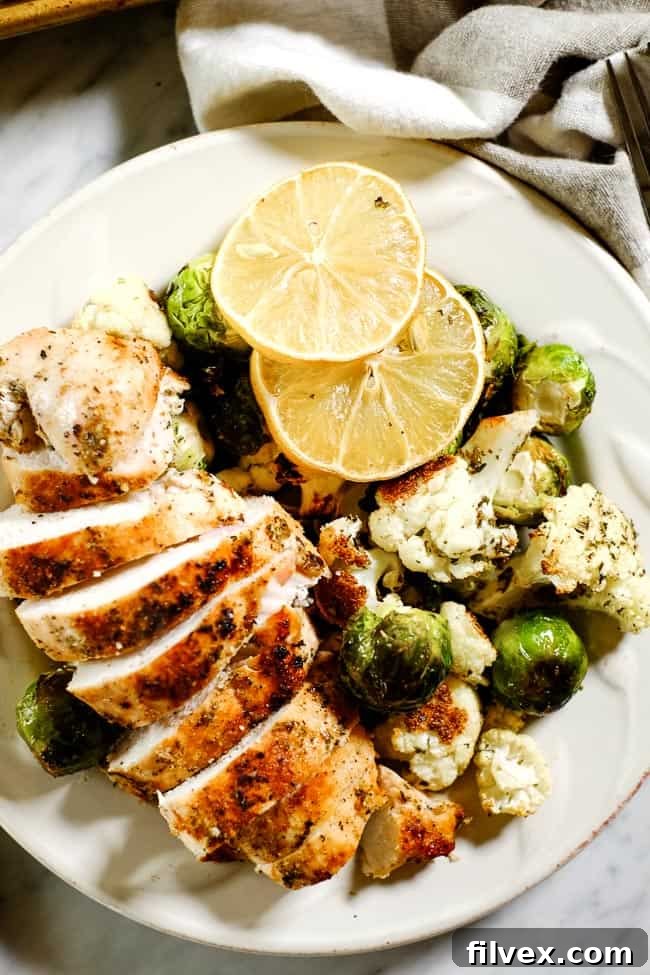 Closer up shot of easy lemon chicken served on a plate with roasted brussels sprouts and cauliflower and garnished with lemon slices. 