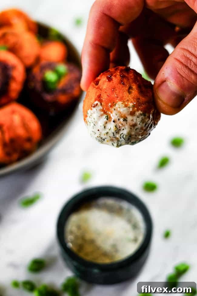 Close-up shot of a buffalo chicken meatball being dipped into a creamy ranch sauce, with a bowl of finished meatballs and green onion garnish in the background.