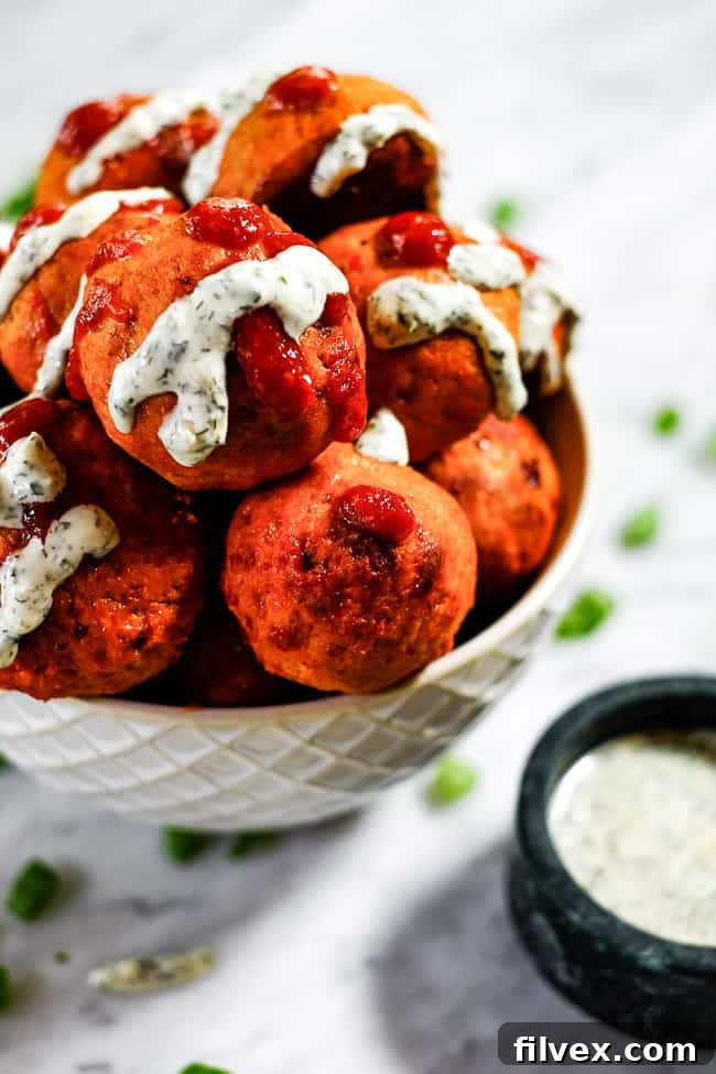Angled shot of buffalo chicken meatballs beautifully arranged in a serving bowl, generously drizzled with ranch and hot sauce. Extra ranch and chopped green onions are served on the side for customization.