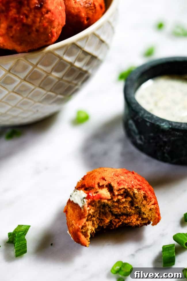 A buffalo chicken meatball with a bite taken out, revealing the moist interior, with creamy ranch dipping sauce and a full bowl of meatballs in the background.