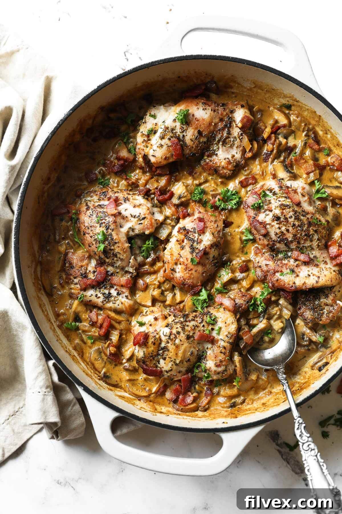 Overhead vertical image of creamy garlic chicken in skillet with a serving spoon dug into dish. Garnished with chopped parsley.