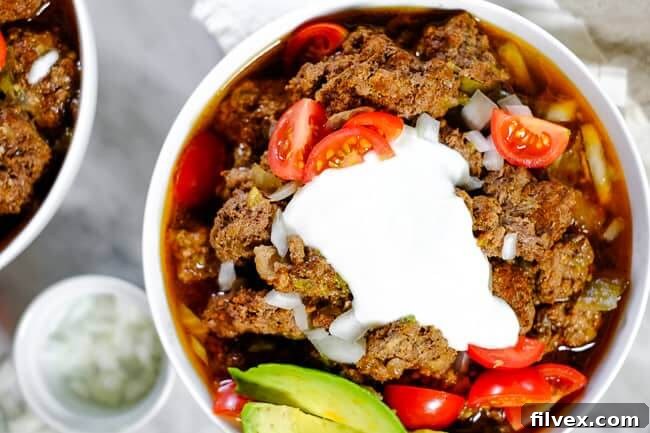 Two bowls of savory Keto chili, generously topped with fresh chopped red onion, vibrant diced tomatoes, creamy slices of avocado, and a dollop of dairy-free coconut yogurt. The rich red chili contrasts beautifully with the colorful toppings.
