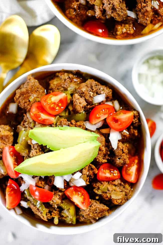 A closer view of a bowl of rich, beefy keto chili, garnished with finely chopped white onion, vibrant red tomatoes, and creamy green avocado slices. The textures and colors are inviting and appetizing.
