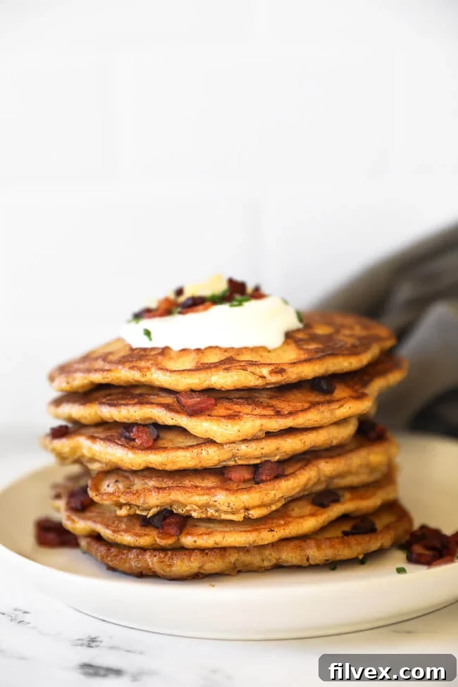 A towering stack of golden bacon pancakes on a clean white plate, artfully topped with a generous dollop of sour cream, crunchy bacon bits, vibrant green chives, and a sprinkle of shredded cheese.