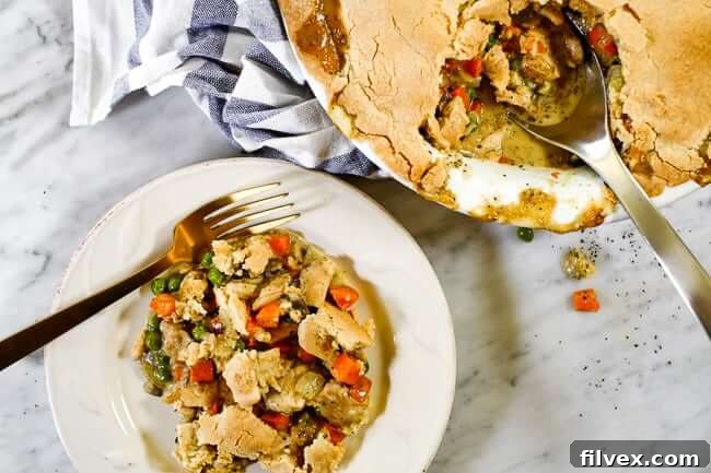 A serving of dairy-free chicken pot pie casserole on a plate with a fork, with the full casserole dish in the background with a blue striped towel and serving spoon.