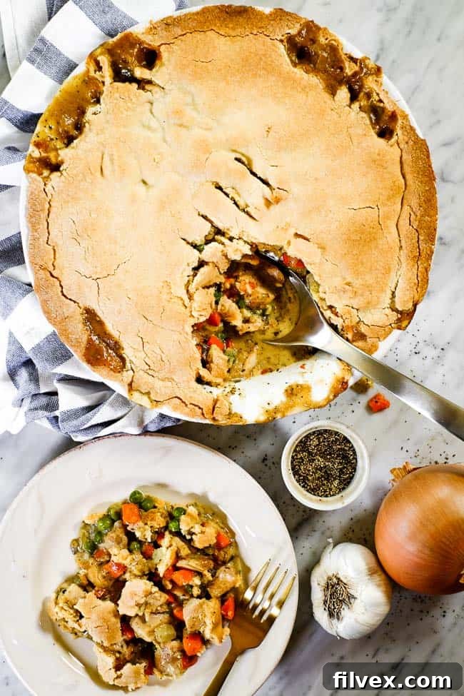 A wider shot of the dairy-free chicken pot pie casserole served on a plate with a fork, with the entire casserole dish in the background with a blue striped towel and serving spoon.