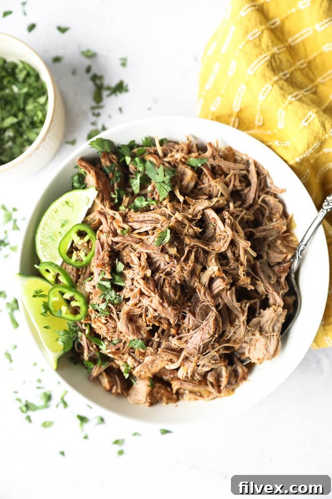 Shredded marinated instant pot mojo pork in a bowl with chopped cilantro, lime wedges and sliced jalapeño on top. 