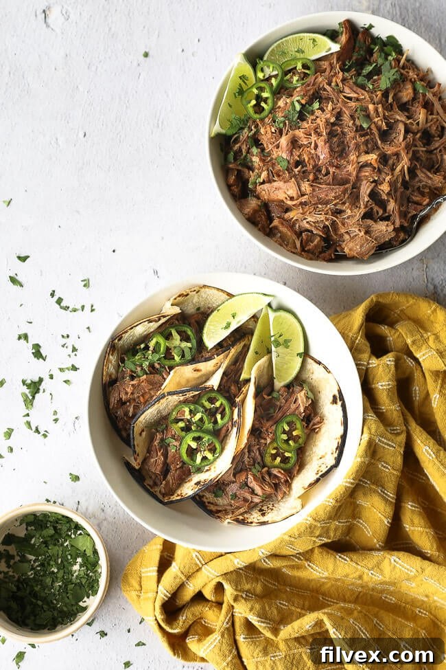 A bowl of shredded mojo pork with some used to make three tacos served up in a dish with lime wedges, chopped cilantro and sliced jalapeño. 