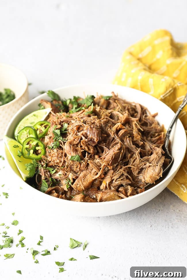 Angled image of marinated mojo pork shredded in a bowl with a serving spoon. Topped with lime wedges, chopped cilantro and sliced jalapeño. 