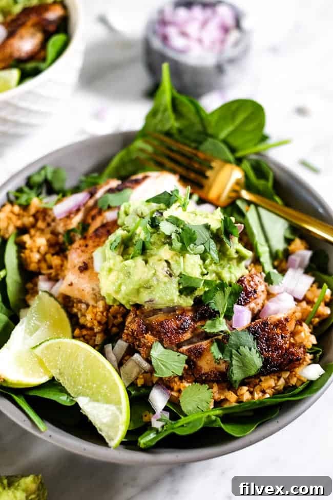 An angled shot showcasing a beautifully arranged chicken burrito bowl, generously topped with vibrant cilantro, creamy guacamole, and finely chopped red onion, with lime wedges adding a pop of color.