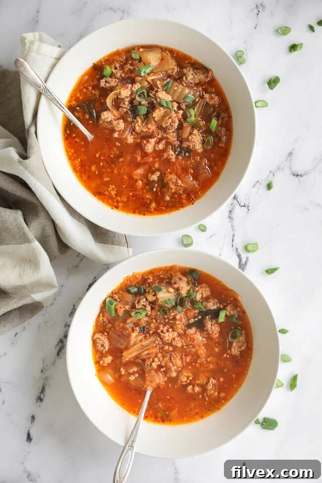Overhead image of two bowls in a vertical line of kimchi soup with chopped green onion on to and spoons dug into the bowls. 