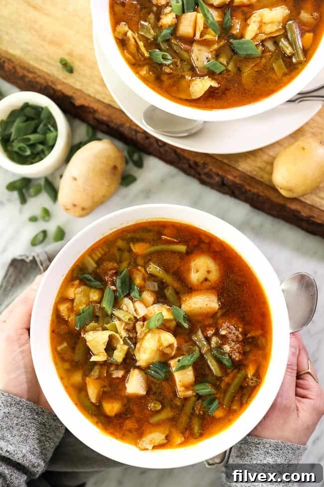 An inviting overhead picture of two bowls of Paleo and Whole30 Hamburger Soup, garnished with green onions and potatoes, with hands reaching for one of the bowls.