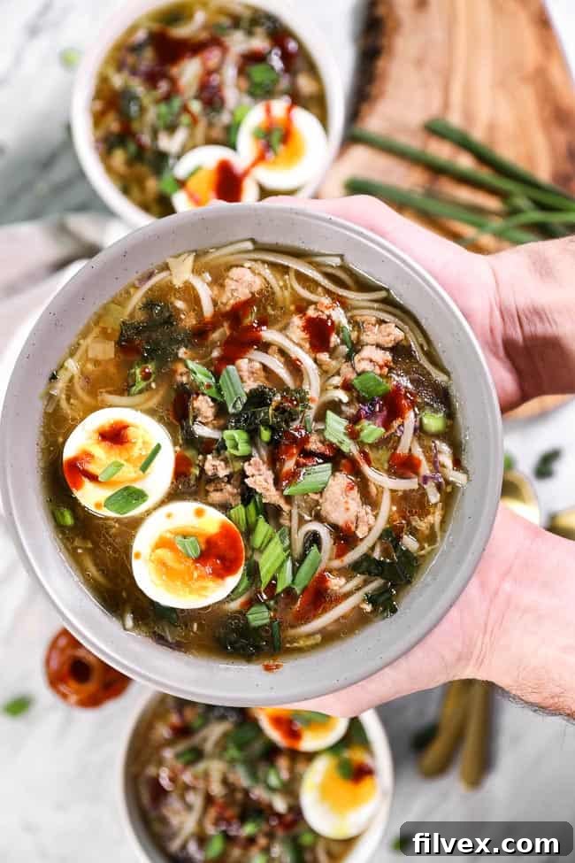 Holding a bowl of pork ramen noodles topped with chopped green onion, a soft boiled egg and hot sauce. 