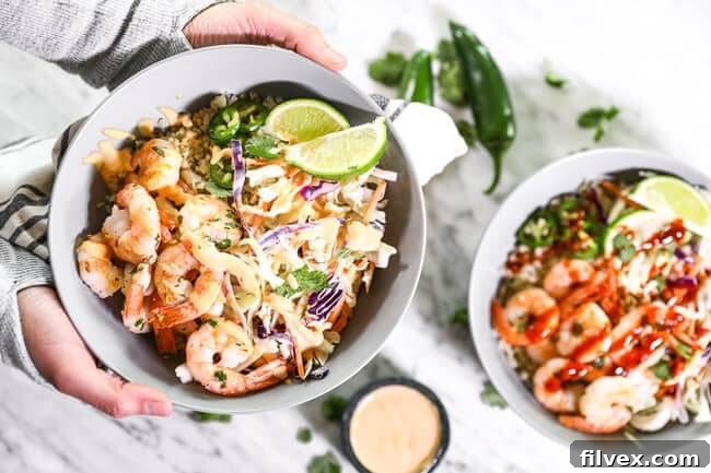 A hand holding a vibrant cilantro lime shrimp bowl, garnished with sauce, jalapeño slices, and lime wedges, against a blurred background of a table with another bowl and sauce. This image showcases the fresh and appealing nature of the dish, highlighting its readiness for a healthy weeknight dinner.