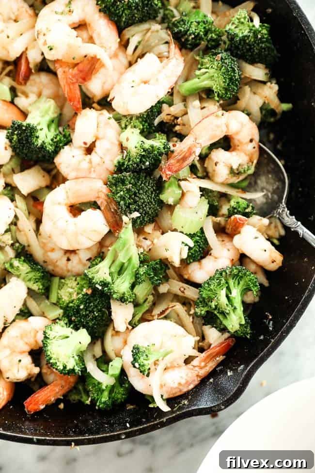 A cast iron skillet filled with a freshly cooked Paleo and Whole30 seafood stir-fry, featuring a tempting medley of shrimp, white fish, vibrant broccoli, crisp celery, and sweet onion, all generously coated in a zesty Cajun seasoning. A serving spoon rests in the pan, highlighting the readiness of the meal.