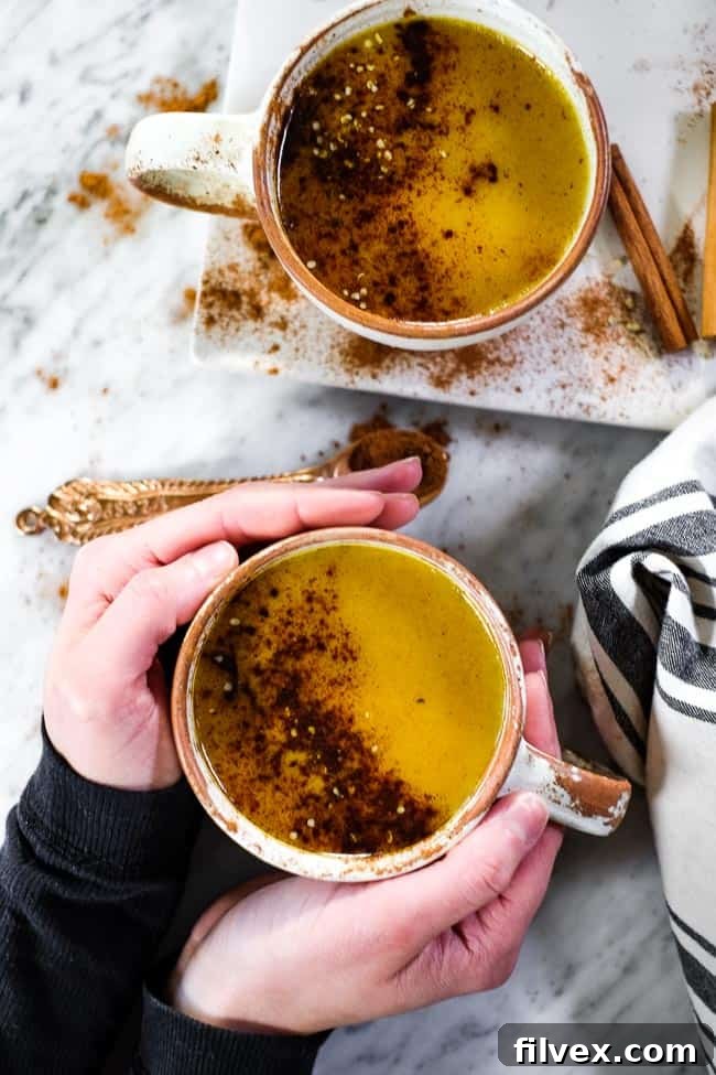 Hands holding a mug of golden milk latte, garnished with cinnamon and hemp seeds. This creamy, cozy, and satisfying caffeine-free turmeric latte is perfect for Paleo, Whole30, Keto, and Vegan lifestyles, making any morning feel complete.