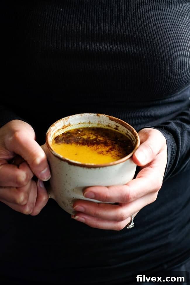 Close-up of a hand holding a single mug of golden milk latte, topped with cinnamon and hemp seeds, emphasizing the creamy texture. This Paleo, Whole30, Keto + Vegan golden milk latte is a satisfying caffeine-free turmeric latte.