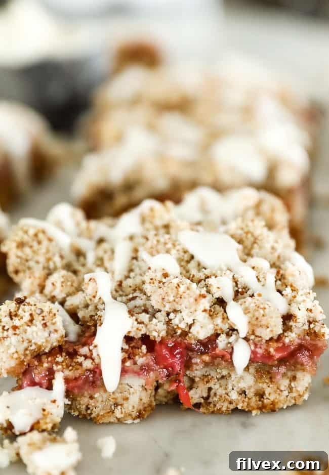 Close-up detailed image of a single strawberry crumble bar, showing the golden-brown, textured crumble topping with a luxurious drizzle of coconut butter melting down its side. The vibrant red strawberry filling is visible beneath the crumble, showcasing the rich layers of this healthy dessert.