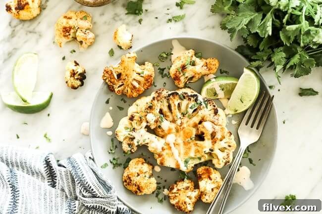 Grilled cauliflower on a plate topped with creamy sauce, chopped cilantro and lime wedges with a fork. 