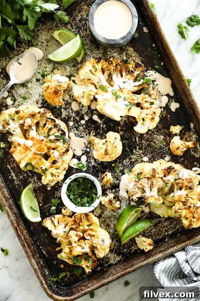 Cauliflower steaks spread out on a baking sheet with cream sauce drizzled on top and topped with cilantro. Cilantro, lime wedges, sauce and a spoon on pan. 