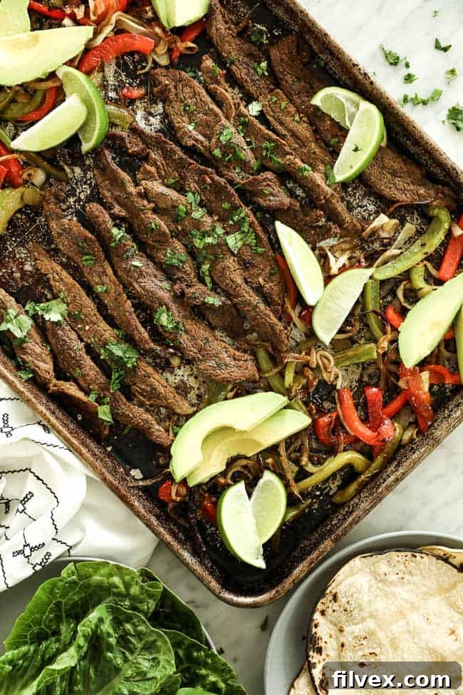 Overhead vertical image of sheet pan fajitas on sheet pan with steak, green and red bell peppers and onion. Topped with fresh cilantro, avocado slices and lime wedges. 