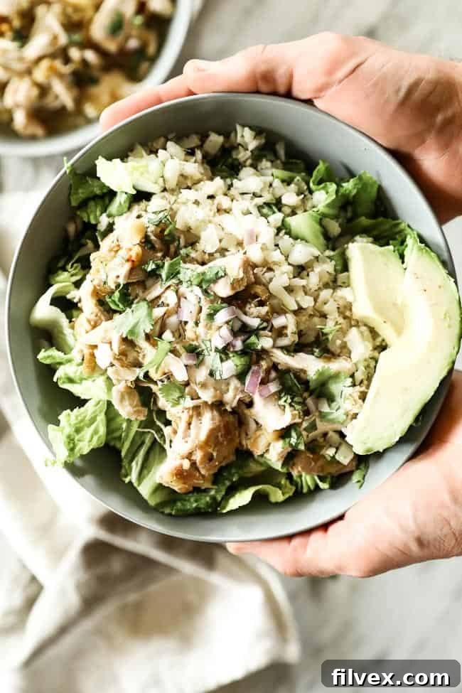 A person holding a healthy bowl of Mexican shredded chicken, featuring a base of romaine lettuce, cauliflower rice, tender shredded chicken, creamy avocado, and a generous topping of fresh chopped red onion and cilantro.